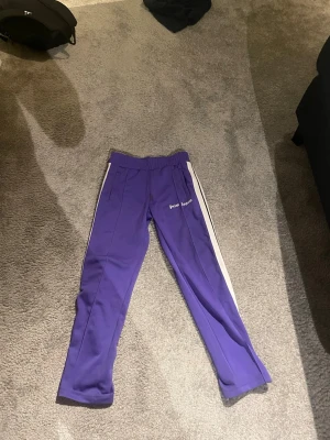 Lila Palm Angels mjukisbyxor M - Säljer ett par lila mjukisbyxor från Palm Angels i storlek M. Byxorna har vita sidoränder, elastisk midja och Palm Angels-logga tryckt på benet. Tillverkade i 100% polyester för en sportig vibe och skön passform.