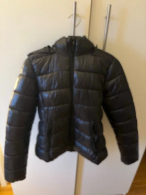 Svart pufferjacka från 157 XS - Säljer en svart pufferjacka från 157 i storlek XS. Jackan har huva, dragkedja framtill och två fickor med dragkedja. Materialet är glansigt och quiltat, perfekt för kyliga dagar. Enkel och stilren design som funkar till det mesta.