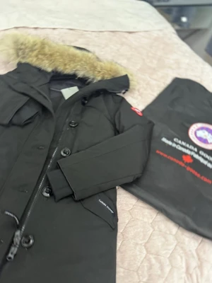 Svart Canada Goose parkas med päls - Säljer en svart parkas från Canada Goose med stor huva och fluffig pälsdetalj. Jackan har dragkedja, stora knappar och praktiska fickor framtill. Klassisk patch på ärmen och lång passform som håller dig varm hela vintern.