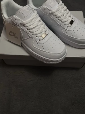 Nike Air Force 1’07 vita sneakers - Nike Air Force 1’07 sneakers i helvit färg med klassisk låg siluett. Skorna har snörning, perforerad tå och platt sula. Tillverkade i skinn med vita detaljer och metallbricka med 'AF1' på snörningen. Ikonisk Nike-logga på sidan och på innersulan.