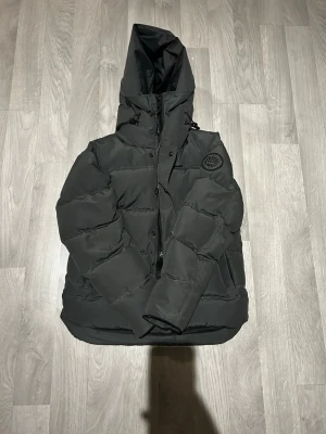 Svart pufferjacka från Canada Goose - Säljer en svart pufferjacka från Canada Goose med huva och diskret logga på ärmen. Lite sönder i insidan Jackan har dragkedja och knappar framtill samt justerbara snören vid huvan. Perfekt för kalla dagar och riktigt snygg till streetwear-stilen.