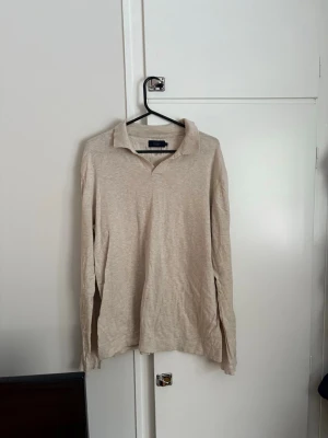 Beige långärmad henley från Riley - Långärmad beige henley från Riley i mjukt och lätt bomullsmaterial. Skjortan har klassisk krage utan knappar och en avslappnad passform. Perfekt för dig som gillar en enkel och clean stil.