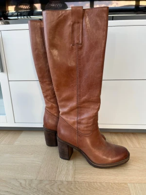Bruna höga stövlar från Rizzo - Snygga bruna stövlar från Rizzo i äkta skinn med rund tå och rejäl klack. Klackhöjd är 7,5 cm. Stövlarna har dragkedja på insidan och en klassisk, tidlös design som passar perfekt till både jeans och kjol. Fodret är svart och sulan har bra grepp. Stövlarna är endast använda vid ett par tillfällen och är som i nyskick. 
