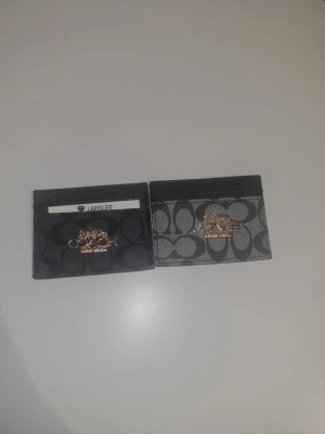 Coach kortfodral i svart och grått - Två stilrena kortfodral från Coach med klassiskt monogrammönster och guldfärgad logga framtill. Ena fodralet är svart och det andra är grått, båda i ett tåligt material som känns lyxigt. Perfekt för att hålla koll på korten i fickan eller väskan.