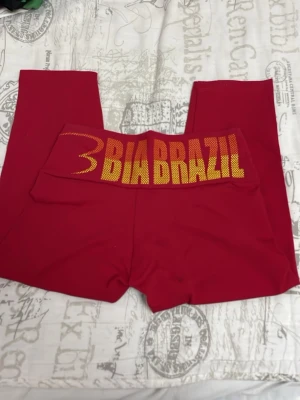 Röda leggings från Bia Brazil - Snygga röda leggings från Bia Brazil med bred midja och gul text baktill. Tillverkade i stretchigt syntetmaterial som passar perfekt för träning. Modellen har en kroppsnära passform och är helfärgade utan mönster.