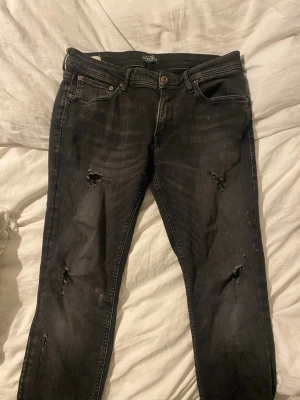 Gråsvarta jeans med fabrikslitningar - Säljer ett par svarta jeans från Jack & Jones i storlek 32/32. Jeansen har skinny fit och fabriksslitningar vilket är väldigt eftertraktat och populärt! Skön passform och inga defekter vilket gör att dem är i mycket bra skick! Pris kan diskuteras via snabb affär 