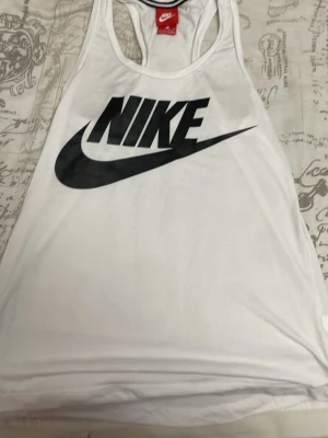Vit linnetopp från Nike - Säljer en vit linnetopp från Nike med stort svart logotryck framtill. Toppen har klassisk racerback och är tillverkad i mjukt och lätt material, perfekt för träning eller chill dagar. Enkel och sportig design som alltid funkar.