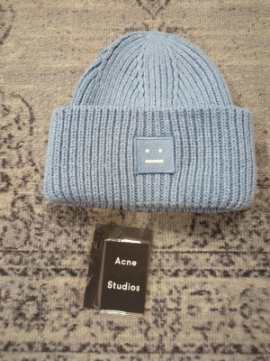 Blå stickad mössa Acne Studios - Säljer en ljusblå ribbstickad mössa från Acne Studios med den klassiska ansiktsloggan framtill. Mössan är i mjuk ullblandning och har bred uppvikt kant för extra stilpoäng. Perfekt accessoar för kyliga dagar.