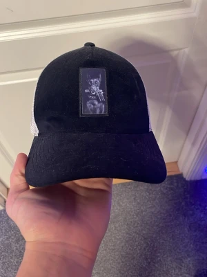 Svart och vit keps från BVALDI - Säljer en snygg svart och vit keps från BVALDI med mesh på sidorna och bak. Framsidan har en patch med en hund och håller i en pistol. Kepsen har justerbar passform och cool streetstyle-vibe.