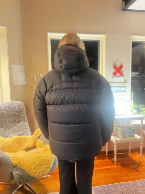 Svart dunjacka från Fjällräven - Säljer en svart dunjacka från Fjällräven med stor huva och klassisk logga på bröstet. Jackan har två stora fickor framtill med tryckknappar och dragkedja. Perfekt för kalla dagar och riktigt skön att bära. Passformen är oversized och jackan är vadderad för extra värme.