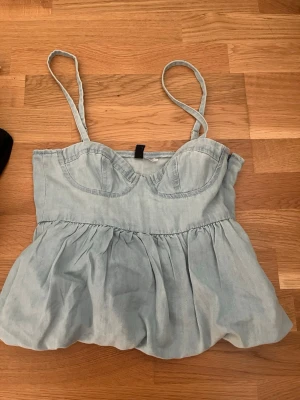 Ljusblå jeanstopp från H&M - Säljer en trendig jeanstopp från H&M i storlek XS som sitter lite för litet för min smak. Toppen har justerbara band, markerade kupor och en volang nedtill. Den har även dragkedja på sidan. Helt oanvänd köpt i spanien