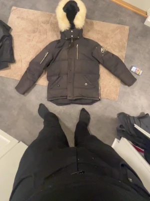 Svart Moose Knuckles dunjacka - Säljer en svart dunjacka från Moose Knuckles med stor huva och beige pälsdetalj. Jackan har flera dragkedjor, logga på ärmen och quiltad design. Perfekt för kalla dagar och har en riktigt schysst streetstil.