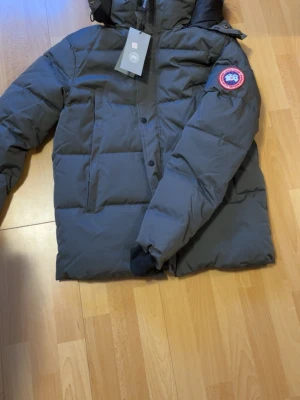 Mörkgrå pufferjacka från Canada Goose Wyndham storlek S - Säljer en mörkgrå pufferjacka från Canada Goose med ikonisk röd och vit patch på ärmen. Jackan har huva, långa ärmar och praktiska fickor framtill. Stängs med både dragkedja och knappar. Perfekt för kalla vinterdagar och riktigt snygg streetstil.