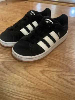 Svarta Adidas Campus sneakers - Säljer ett par klassiska Adidas Campus sneakers i svart mocka med vita ränder och Ortholite-innersula. Skorna har svart snörning, vit sula och den ikoniska Adidas-loggan på plösen. Perfekta för dig som gillar retrostil och streetwear.