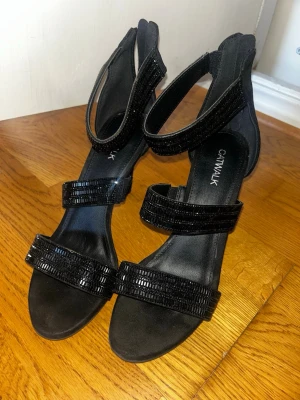 Svarta sandaler med klack från Catwalk - Snygga svarta sandaler från Catwalk med breda remmar dekorerade med svarta pärlor och paljetter. Skorna har öppen tå, klack och rundad form. Materialet är mocka-liknande syntet som ger en lyxig känsla. Perfekta för dig som vill ha något extra till outfiten.