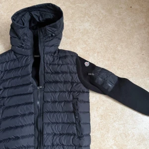 Svart pufferjacka från Moncler - Svart pufferjacka från Moncler med stickade ärmar och huva med snörning. Jackan har dragkedja framtill, två fickor med knappstängning och en extra ficka med dragkedja på ärmen. Snyggt Moncler-märke på armen och ribbade muddar vid ärmslut och nederkant.