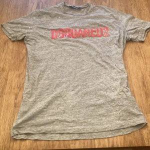 Grå DSQUARED2 t-shirt med röd logga - Grå t-shirt från DSQUARED2 i storlek L med röd logotyp framtill. Klassisk passform och korta ärmar. Tillverkad i mjuk bomull, perfekt för en avslappnad och trendig look.