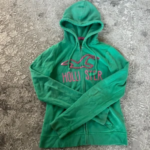 Grön hoodie från Hollister med dragkedja - Snygg grön hoodie från Hollister i storlek XS med rosa broderad logga och text på bröstet. Tröjan har huva med snörning, långa ärmar och praktiska fickor framtill. Tillverkad i mjuk bomullsmix och stängs med dragkedja. Perfekt för chill dagar.