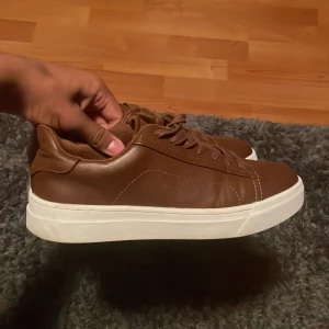Bruna calvin Klein sneakers med vit sula - Snygga bruna calvin Klein sneakers med slät ovandel i skinn och klassisk vit platt sula. Skorna har rund tå och snörning framtill för en clean look. Perfekta för dig som gillar stilrena och enkla sneakers med lite extra edge.