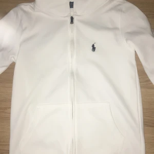 Vit hoodie från Polo Ralph Lauren - Vit hoodie från Polo Ralph Lauren med klassisk logga broderad på bröstet. Tröjan har dragkedja framtill, snörning i huvan och en stor magficka. Perfekt för dig som gillar stilrena och sportiga plagg med premiumkänsla. Koftan känns lite mindre is storlek, skulle säga mellan S och XS, men annars är en toppen