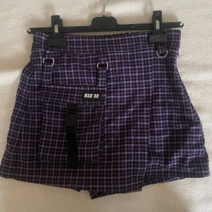 Lila-rutiga shorts från Bershka - Säljer ett par lila och svarta rutiga shorts från BSK med cool utility-look. De har en stor ficka framtill med spänne och metallringar upptill. 
