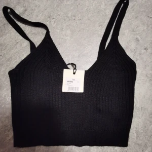 Svart ribbad linnetopp från Bik Bok - Svart ribbad linnetopp från Bik Bok med smala axelband och v-ringad hals. Toppen är croppad och har en tight passform som framhäver siluetten. Perfekt att styla med höga jeans eller kjol för en trendig look.