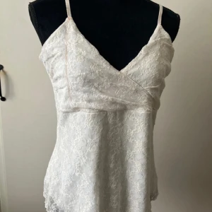 Vit spetstopp från KappAhl - Supersöt vit glittrig spetstopp från KappAhl med tunna axelband. Den är täckt av spets med glittrig blommigt mönster och har en feminin v-ringad hals. Perfekt för dig som gillar romantisk och fräsch stil. I super bra skick!