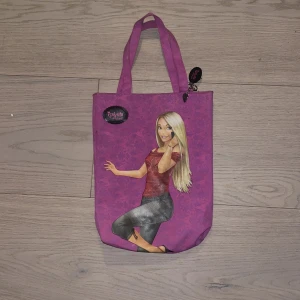 Isabella Fiorella rosa barnshoppingväska - Säljer en rosa barnshoppingväska med tryck av en tecknad tjej på framsidan. Väskan har två handtag och ett litet hängsmycke med hjärtan. Materialet är troligtvis polyester och väskan har en mjuk, lätt form. Perfekt för att bära böcker eller småsaker.