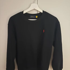 Svart tröja från Polo Ralph Lauren - Svart tröja från Polo Ralph Lauren med klassisk rund halsringning och rött broderat logomärke på bröstet. Tröjan har lång ärm och är tillverkad i mjuk bomull, perfekt för en clean och stilren look.