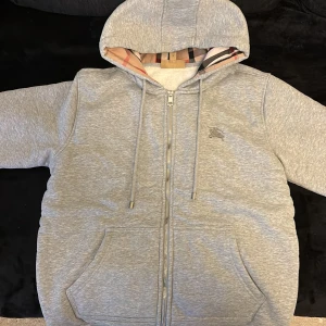Grå hoodie från Burberry med dragkedja - Snygg grå hoodie från Burberry med klassiskt rutigt foder i huvan och insidan. Tröjan har dragkedja, snörning vid huvan och två stora fickor fram. Broderad Burberry-logga på bröstet. Perfekt för dig som gillar exklusiva streetwear-vibes.