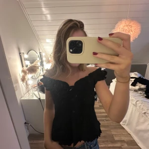 Svart blus med volang och knappar - Säljer en svart blus med offshoulder och volangdetaljer längs halsringning och nederkant. Blusen har knappar framtill och en lätt rynkad effekt vid bysten. Materialet känns som bomull eller polyester och toppen har en feminin, croppad passform.