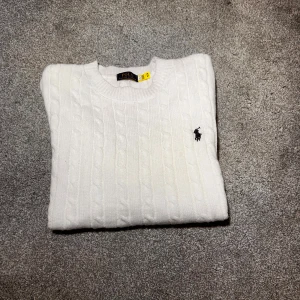 Stickad tröja Polo Ralph Lauren - Snygg vit kabelstickad tröja från Polo Ralph Lauren med klassisk rund hals och broderad logga på bröstet. Tröjan har långärmad passform och ribbade muddar vid ärmslut och nederkant. Perfekt för dig som gillar stilren och tidlös design.