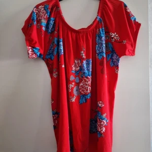 Röd blommig topp  Miss Midas - Röd topp från Miss Midas med färgglada blommor i blått och vitt.. Toppen har korta ärmar, rundad halsringning med resår och en luftig passform. Perfekt för varma dagar och ger en färgstark look.