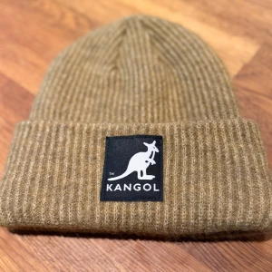 Ribbad beige mössa Kangol x H&M - Snygg ribbstickad mössa i beige från Kangol x H&M. Klassisk design med uppvikt kant och ikonisk kanguru-logga framtill. Perfekt för att hålla värmen och ge din look en chill vibe. Materialet är mjukt och skönt, passar till både street och casual outfits.