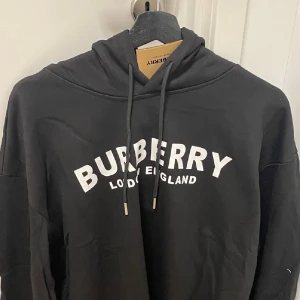 Svart hoodie från Burberry med tryck - Svart hoodie från Burberry med vit text 'BURBERRY LONDON ENGLAND' på bröstet. Tröjan har huva med snörning och en avslappnad passform. Perfekt för dig som vill ha en stilren och lyxig streetwear-look.