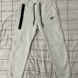 Grå mjukisbyxor från Nike, storlek M - Snygga ljusgrå mjukisbyxor från Nike med svart logga på vänster lår och svarta snören i midjan. Byxorna har en lång svart dragkedja på sidan och är tillverkade i en mjuk blandning av bomull och polyester. Perfekt för chill eller träning.