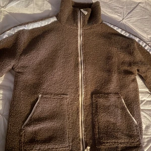 Brun teddyjacka med vita detaljer - Mysig brun teddyjacka med dragkedja framtill och två stora fickor. Jackan har vita ränder längs ärmarna och hög krage som ger en cool och avslappnad vibe. Perfekt för kyliga dagar när du vill vara både varm och snygg.
