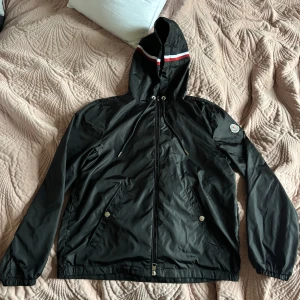 Svart vindjacka från Moncler - Svart vindjacka från Moncler med huva och dragkedja. Jackan har vita och röda ränder på huvan, två fickor med tryckknappar och Moncler-logga på ärmen. Tillverkad i lätt polyester, perfekt för blåsiga dagar. Sitter perfekt på nån som är runt 170. Snygg och Grisch!
