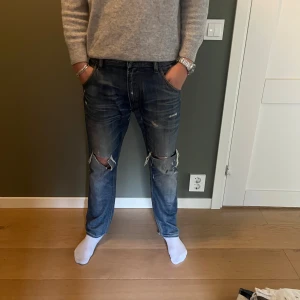 Blå slitna jeans från Diesel - Säljer ett par blå jeans från Diesel med snygga slitningar och stora hål vid knäna. Jeansen har klassisk femficksdesign, raka ben och orangea sömmar. Materialet är jeans i bomull och passformen är normal. Perfekta för dig som gillar en avslappnad och trendig look.