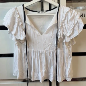 Vit blus med volangärmar från Zara - Supersöt vit blus från Zara med volangärmar och spetsdetaljer. Blusen har v-ringning, svarta knytband och en lös, flowy passform. Perfekt för dig som gillar romantisk och feminin stil.