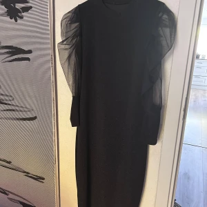 Svart glittrig klänning med puffärm  - Svart klänning med glittrigt tyg och transparenta puffärmar i mesh. Klänningen har en rak siluett och rund halsringning. Perfekt för dig som vill sticka ut med en elegant och trendig look.