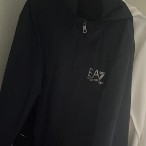 Svart vindjacka EA7 Emporio Armani - Svart vindjacka från EA7 Emporio Armani med huva och dragkedja framtill. Jackan har stilren design med logotyp på bröstet och meshfoder på insidan. Perfekt för dig som gillar sportig och clean look.