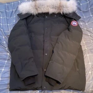 Svart dunjacka från Canada Goose - Säljer en svart dunjacka från Canada Goose med avtagbar huva och fluffig pälsdetalj. Jackan har lång ärm, dragkedja och knappar framtill samt klassisk logopatch på ärmen. Perfekt för kalla vinterdagar och har en snygg, stilren look.