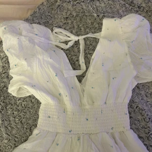 Vit blommig klänning med puffärm - Superfin vit klänning från NA-KD med små blå blommor, puffärmar och smock i midjan. Djup v-ringning med knyt i ryggen och lätt, luftig bomullskänsla. Perfekt för varma dagar och ger en romantisk vibe.