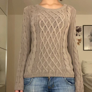 Beige stickad tröja med kabelmönster - Snygg beige stickad tröja med klassiskt kabelmönster framtill och längs ärmarna. Rund halsringning och långärmad modell som sitter normalt. Perfekt för dig som gillar en chill och stilren look med lite extra struktur i materialet.