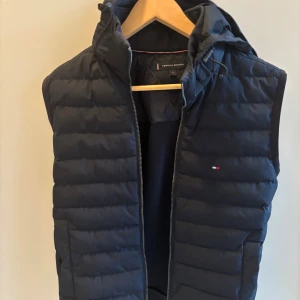Mörkblå dunväst från Tommy Hilfiger - Snygg mörkblå dunväst från Tommy Hilfiger med huva och dragkedja framtill. Västen har quiltad design och diskret logga på bröstet. Perfekt för lager-på-lager och har en sportig vibe med praktiska fickor och justerbar huva.