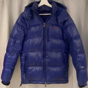 Blå dunjacka Polo Ralph Lauren - Snygg blå dunjacka från Polo Ralph Lauren i storlek XS men sitter mer som S. Jackan har huva, flera dragkedjefickor och ett rött Polo-logo på bröstet. Puffad design med lång ärm och quiltade paneler. Perfekt för kalla dagar och har en modern, sportig look. Jackan har tappat lite glans inre underarmen och har ett litet hål som man ser på bilderna men detta är saker man såklart inte tänker på.