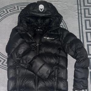 Svart dunjacka från Sail Racing - Svart dunjacka från Sail Racing med huva och dragkedja. Jackan har en puffig design, logga på bröstet och på huvan, samt justerbara muddar. Perfekt för kalla dagar och riktigt snygg streetstil.