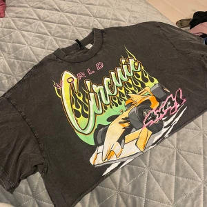 Svart croppad t-shirt Divided XS - Cool svart croppad t-shirt från Divided med färgglatt racingtryck och texten 'World Circuit 4x4'. T-shirten har korta ärmar, rund hals och är gjord i mjuk bomull. Perfekt för dig som gillar streetstyle och sportiga vibes.