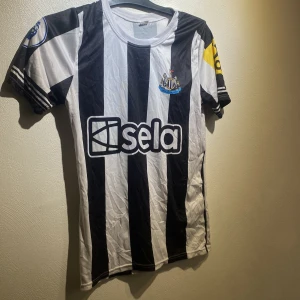 Newcastle United Isak 14 matchtröja - Säljer en svartvit randig Newcastle United fotbollströja med Isak 14 på ryggen. Tröjan har klubbmärke på bröstet, Premier League-märke på ärmen och sponsorlogga framtill. Kortärmad modell i lätt och ventilerande material, perfekt för match eller träning. Den är lite skrynlig men det är bara o platta.